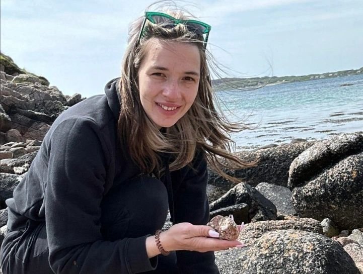 Luisa Hendry - Scottish Geologist, rock in hand, knelt in the foreground beside rocks with the sea in the background